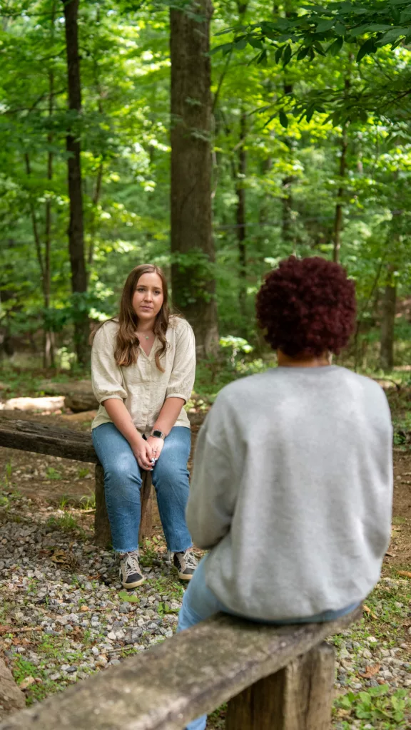 A teen and counselor talk on logs in the woods during outdoor therapy